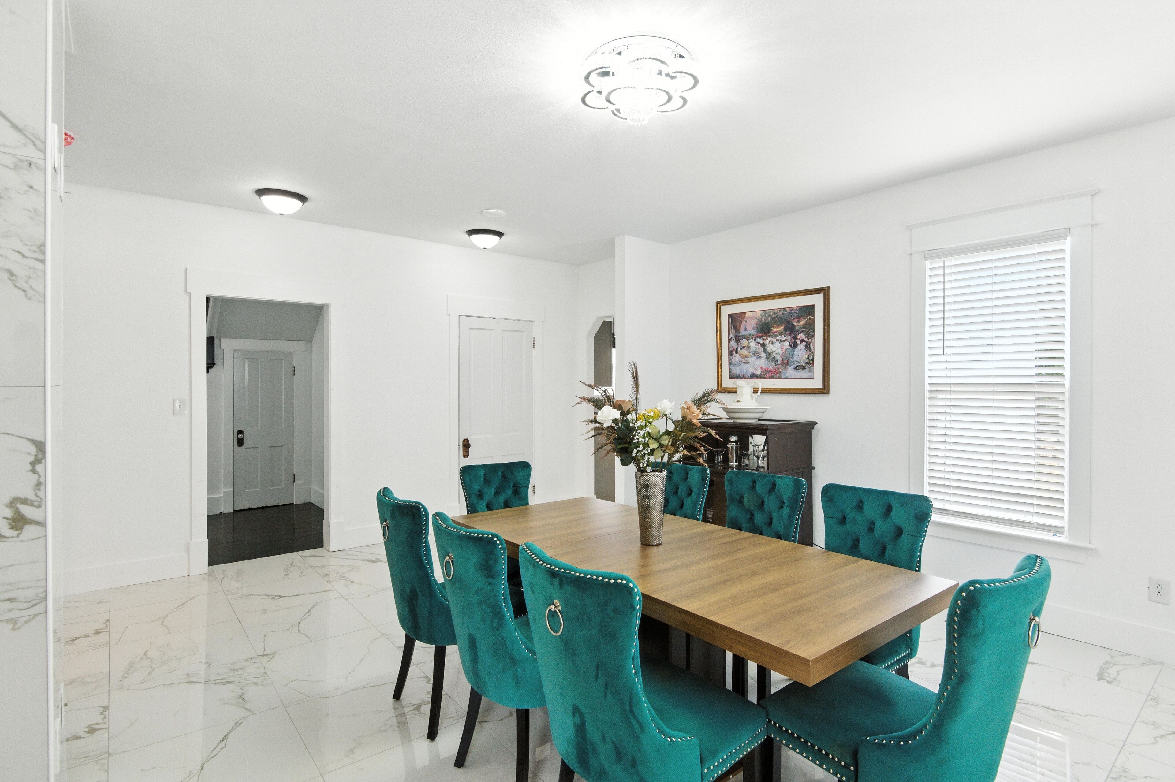 A dining room with a wooden table and teal upholstered chairs, white walls, and a ceiling light fixture.