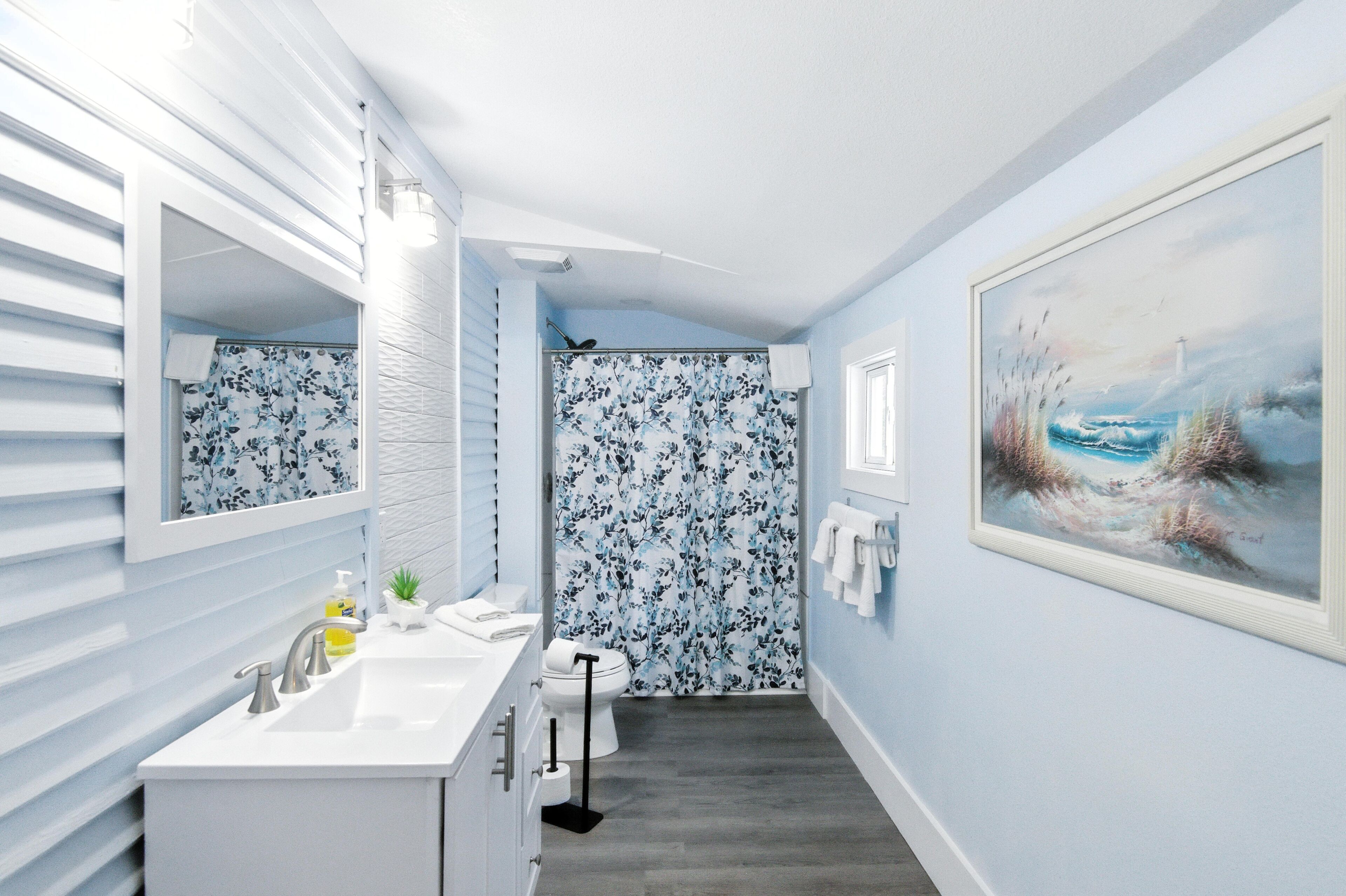A bathroom with a white vanity, a shower with floral wallpaper, and a painting of a seascape on the wall.