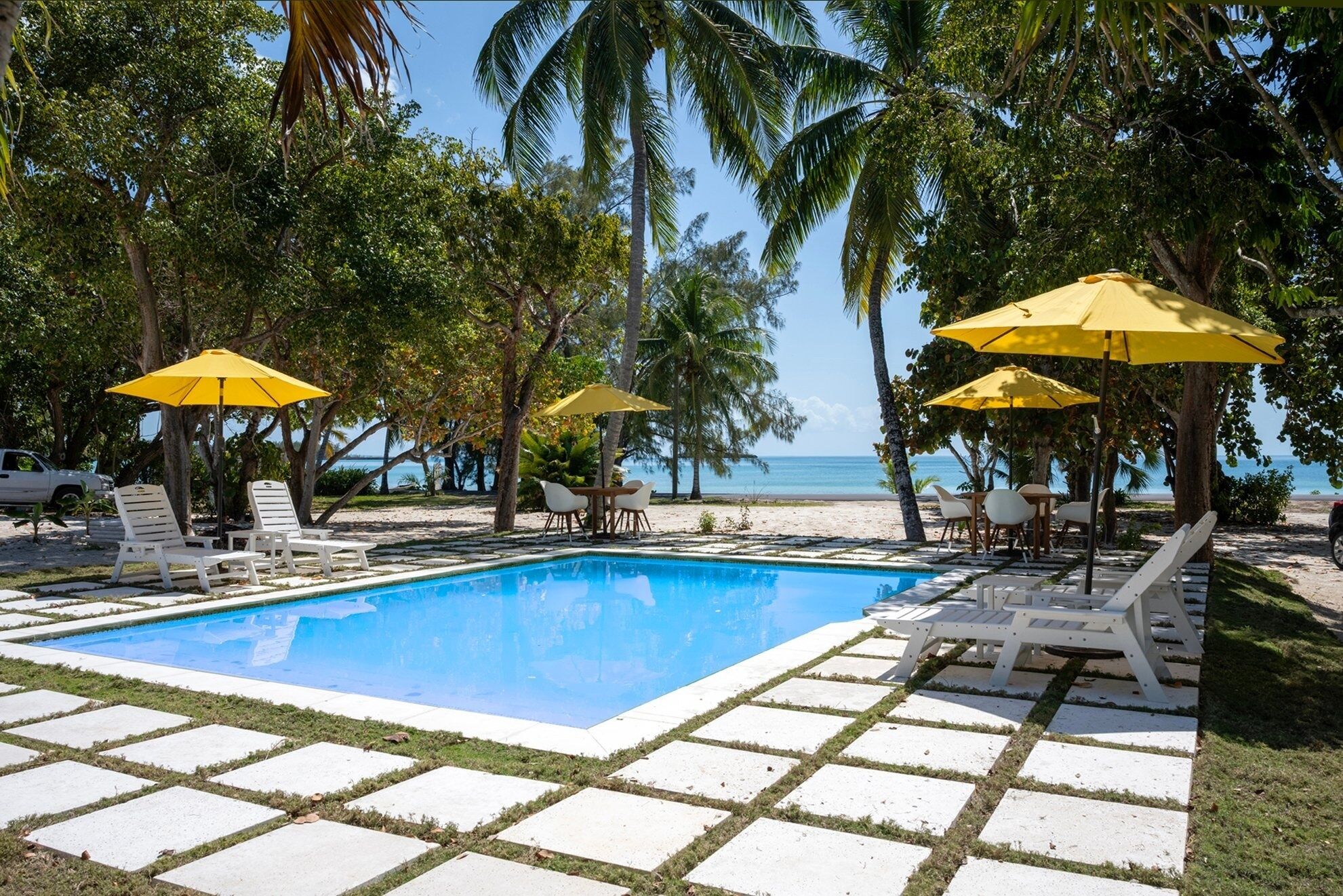 A pool area with white lounge chairs and yellow umbrellas, surrounded by palm trees and a view of the beach.