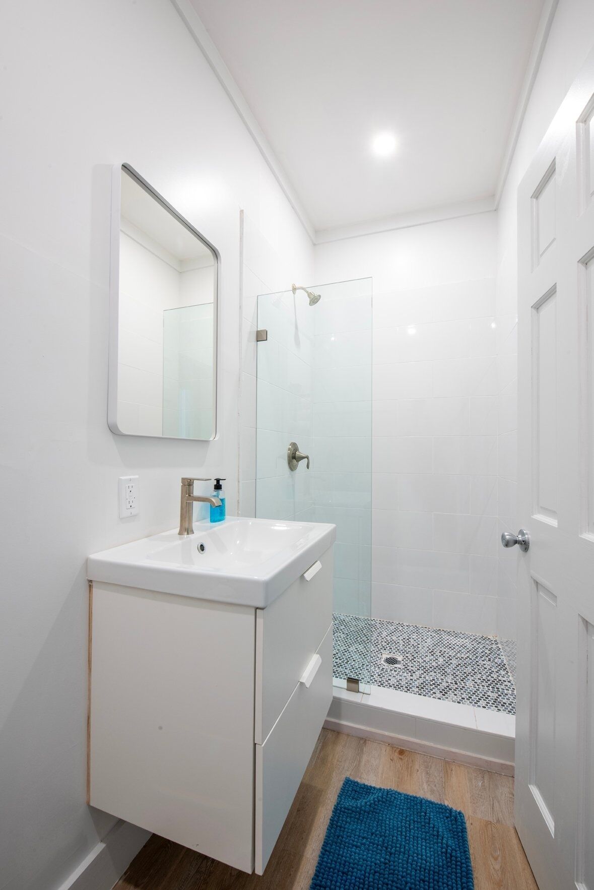 A modern bathroom with a glass shower enclosure, a white sink with a soap dispenser, and a blue rug on the floor.