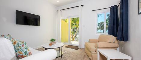 Una sala de estar moderna con un sofá blanco, un sillón color beige, una mesa de centro y un televisor montado en la pared. Se ve una ventana con cortinas, un ventilador de techo y una vista a una pared amarilla y follaje en el exterior.