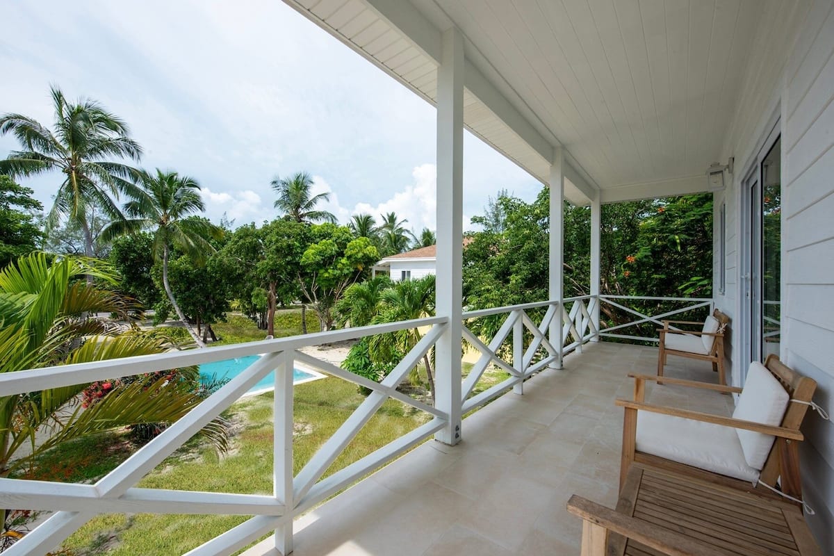 A covered balcony with a pool, wooden chairs, and lush greenery.