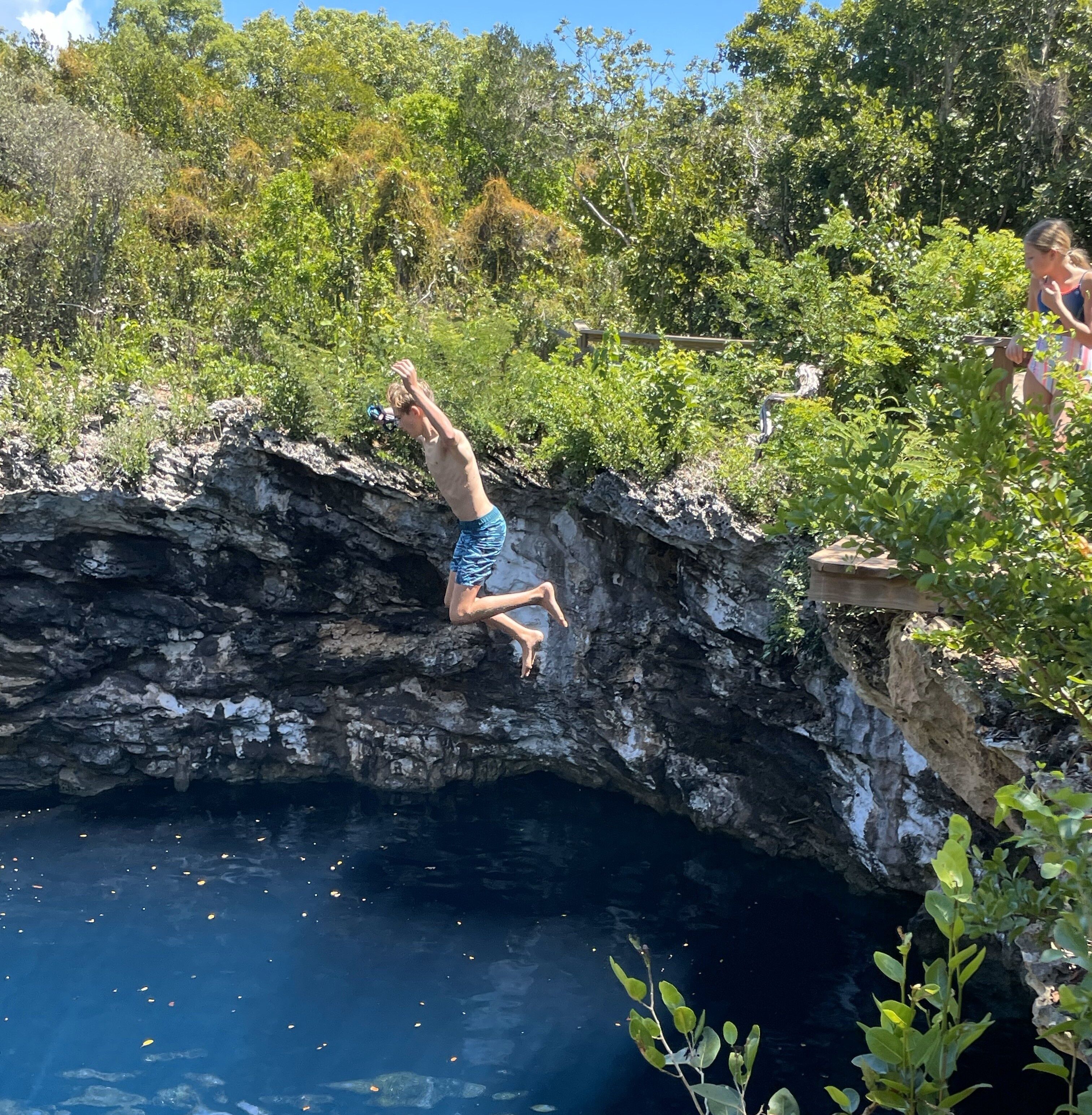 Eine Person springt von einer felsigen Klippe in ein Gewässer, umgeben von viel Grün.