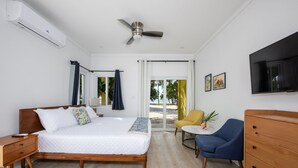 A hotel room with a bed, a desk, a chair, a TV, and a view of the beach. - Laughing Bird Studio #7 - Brand New Stylish Apartment, Pool, Bea 1 Bedroom Condo (Governor's Harbour)