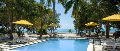 A pool area with white lounge chairs and yellow umbrellas, surrounded by palm trees and a view of the ocean.