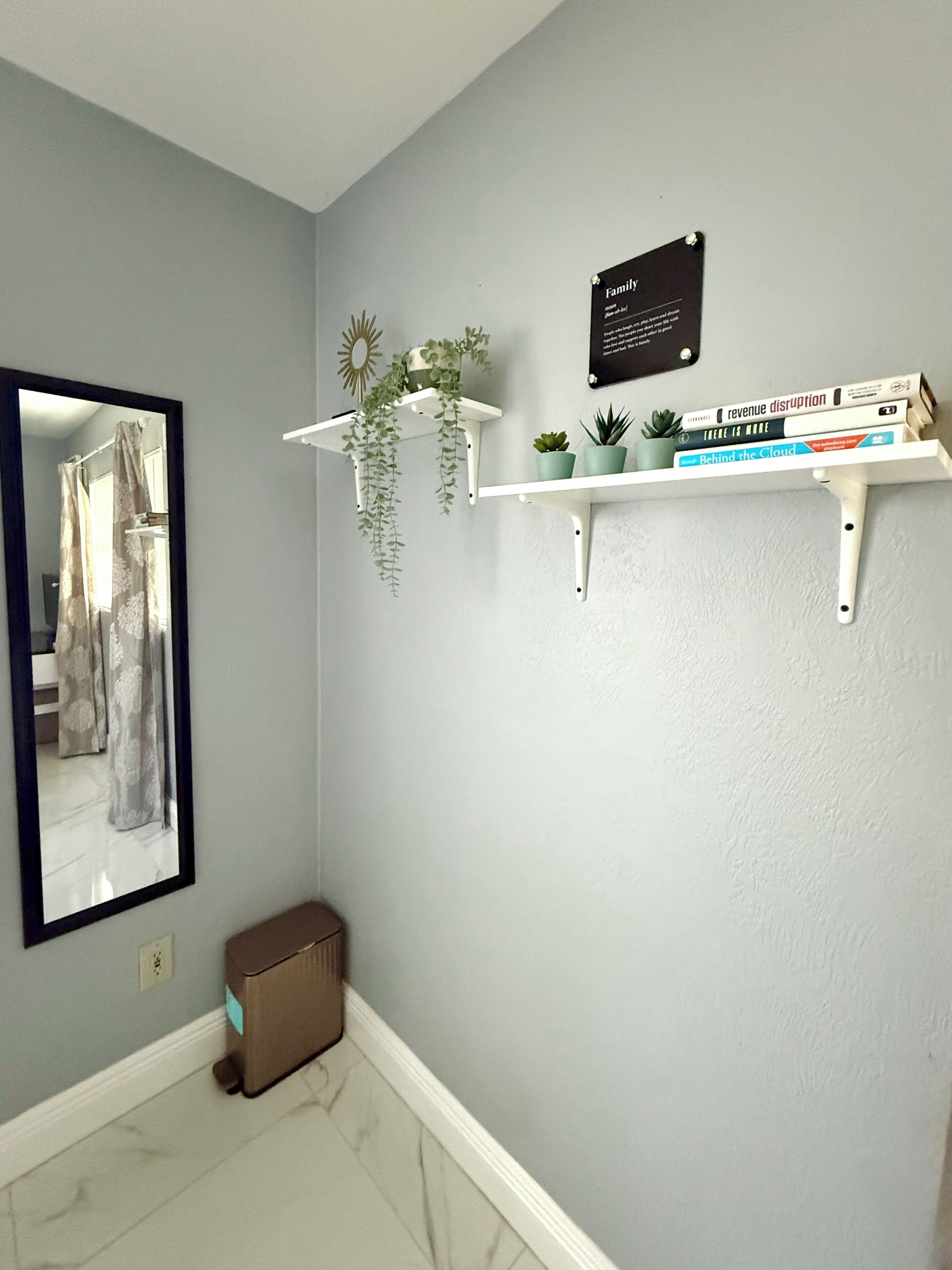 Ein Zimmer mit hellblauen Wänden, einem weißen Boden, einem Spiegel, einer kleinen Mülltonne und zwei weißen Regalen mit Pflanzen und Büchern.
