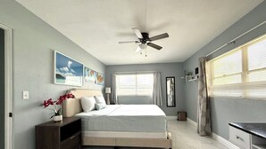 Une chambre à coucher comprenant un lit, un ventilateur de plafond, une fenêtre avec des rideaux, une commode et un bureau avec une chaise.