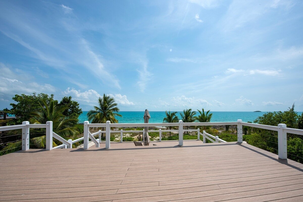 A wooden deck with white railings overlooking a beach with palm trees and the ocean.