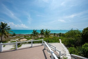 A wooden deck with white railings overlooking a beach with palm trees and a clear blue sky. - Ware-a-dise - Newly Remodeled Home On Calm Caribbean 3 Bedroom Home (South Palmetto Point)