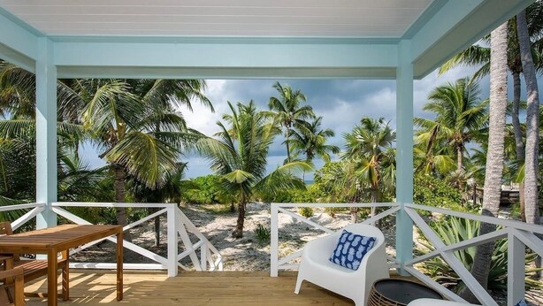 Ein überdachter Außenbereich mit Holzterrasse, weißer Möbel und Blick auf Palmen und Strand.