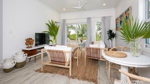Ein modernes Wohnzimmer mit Fernseher, zwei Sofas, einem Esstisch und großen Fenstern mit Vorhängen.