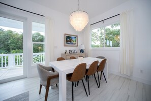 A dining room with a white table, brown chairs, a chandelier, and a view of greenery through large windows. - Silver Palms - Brand New Designer Home On Banks Road 2 Bedroom Home (North Palmetto Point)