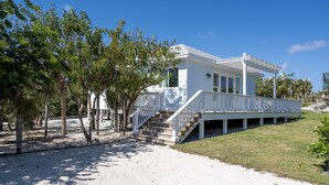 Una casa blanca frente a la playa, con un porche cubierto y escaleras que llevan a la entrada.