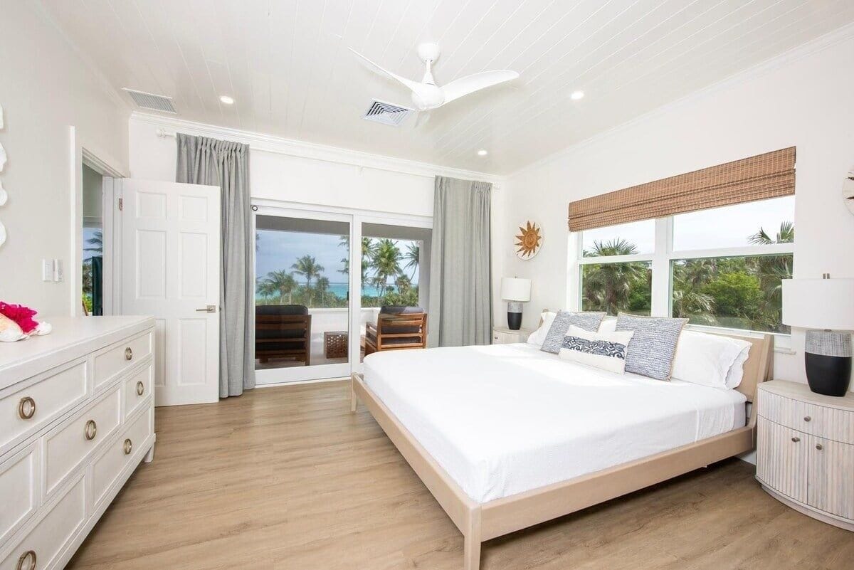 A bedroom with a large bed, a ceiling fan, a window with a view of palm trees, and a sliding glass door leading to an outdoor area.