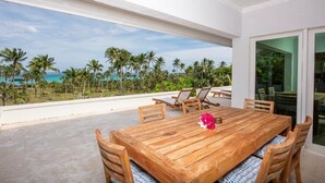 A wooden dining table with chairs, a flower centerpiece, and a view of the ocean and palm trees.