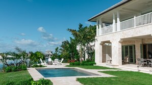 Un área junto a la piscina con vista despejada al océano, un porche cubierto con mobiliario exterior y abundante vegetación.