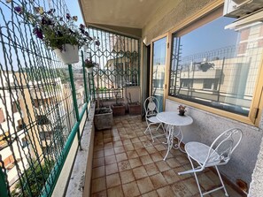 A balcony with a tiled floor, two white metal chairs, a small round table, and a potted plant.