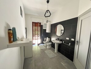 A modern bathroom with a white vanity, a round mirror, a pedestal sink, and black tiled walls. - Spacious 3 Bedrooms with Balcony&AC in Vaticano (Roma)