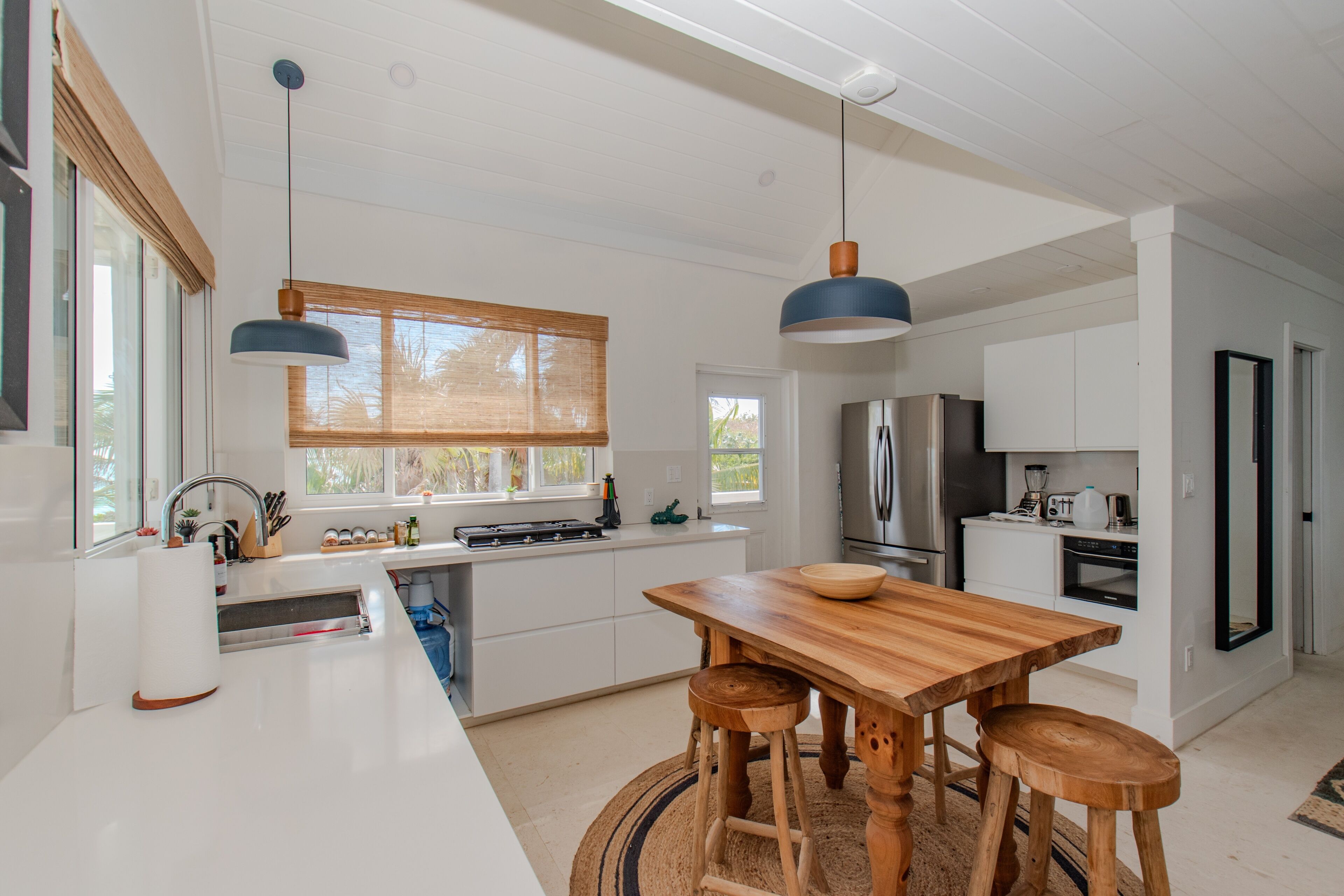 A modern kitchen with white cabinetry, a wooden dining table, two bar stools, a stainless steel refrigerator, and a built-in oven.