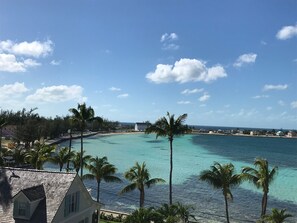 A coastal view with turquoise waters, palm trees, and a white building with a sloped roof. - Sky Beach Club Bungalow 3 Steps From Beach W/ Club, Pool 1 Bedroom Bungalow (Governor's Harbour)
