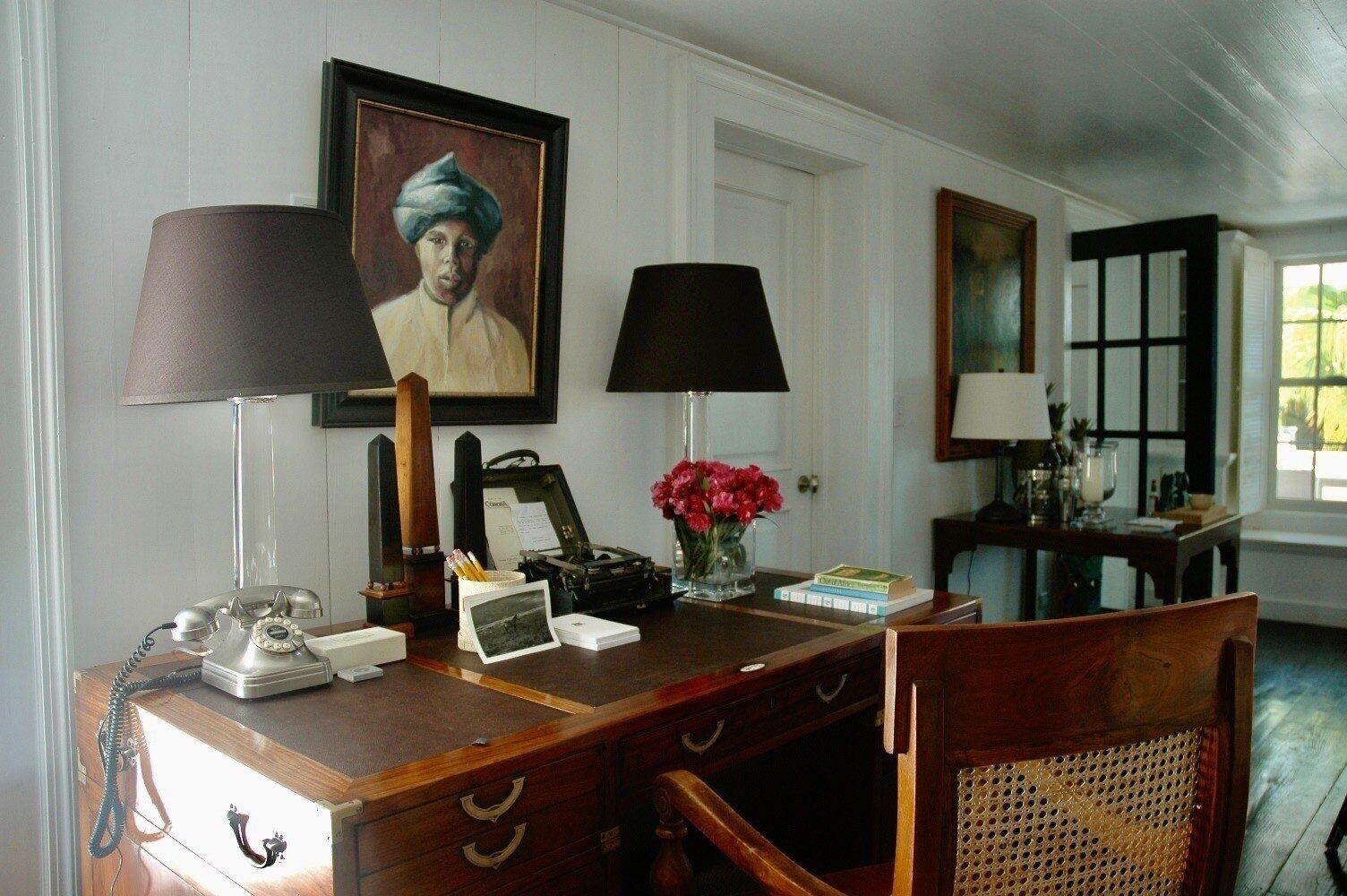 A room with a wooden desk, two lamps, a telephone, a typewriter, a vase with flowers, and framed pictures on the wall.