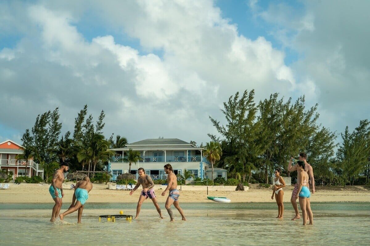 Une scène de plage où l’on voit des gens jouer dans l’eau, une maison en arrière-plan et des palmiers.