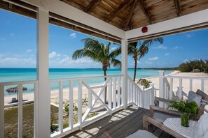 Una terraza de madera con barandas blancas que da a una playa de aguas turquesas y palmeras.