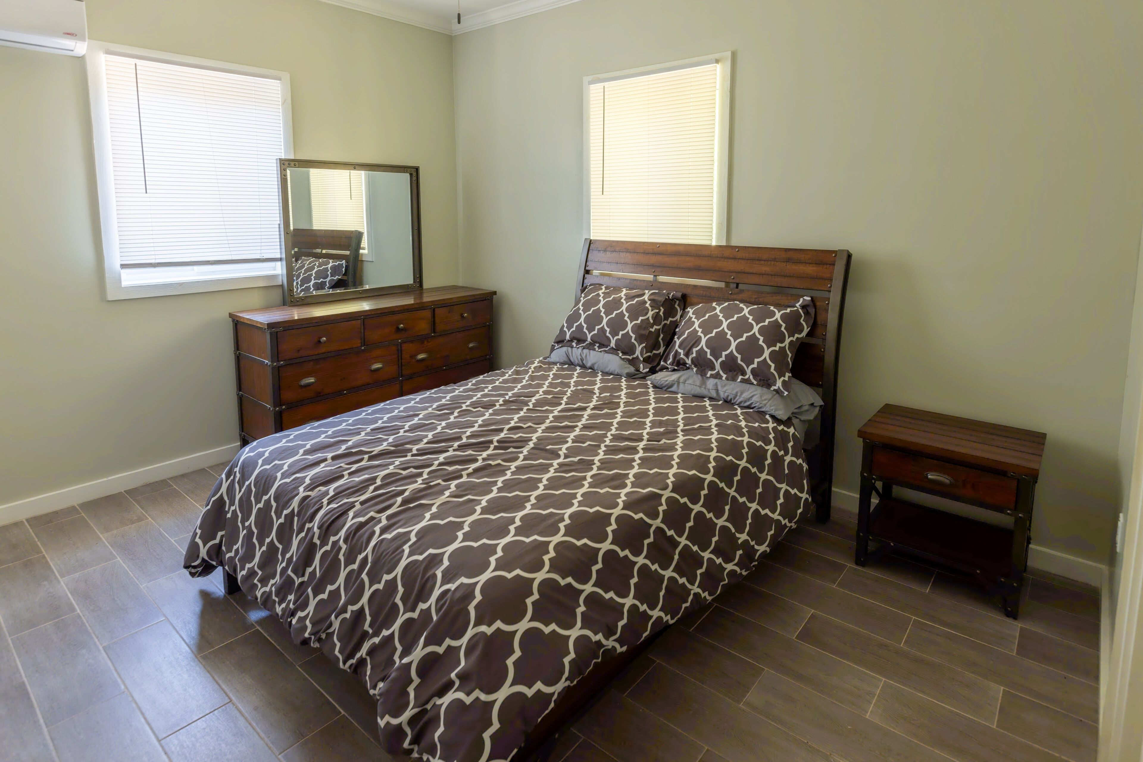 A bedroom with a bed, dresser, nightstand, and two windows with blinds.