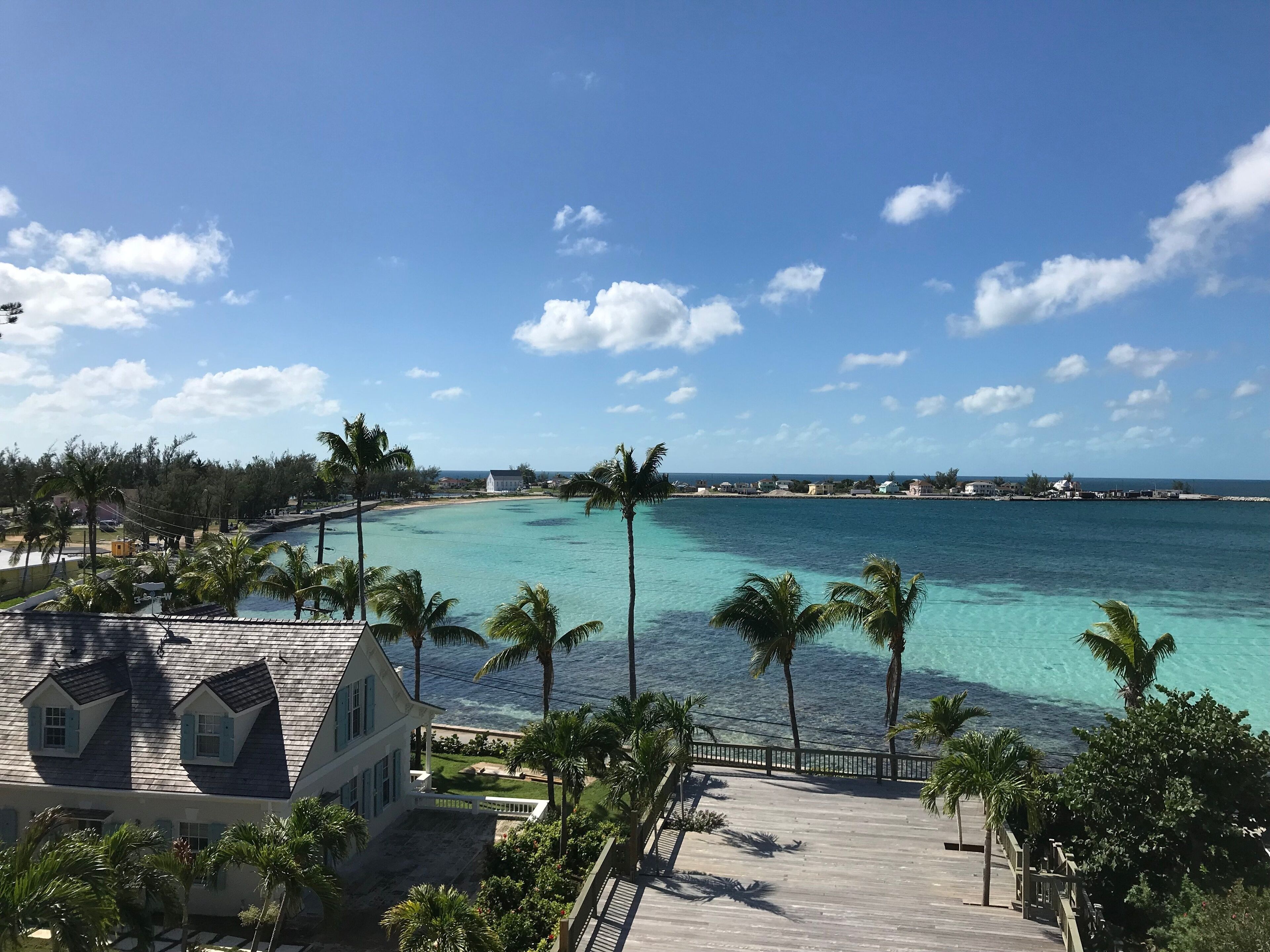 Un resort con una terraza de madera que da a aguas turquesas, rodeado de palmeras y un cielo despejado.