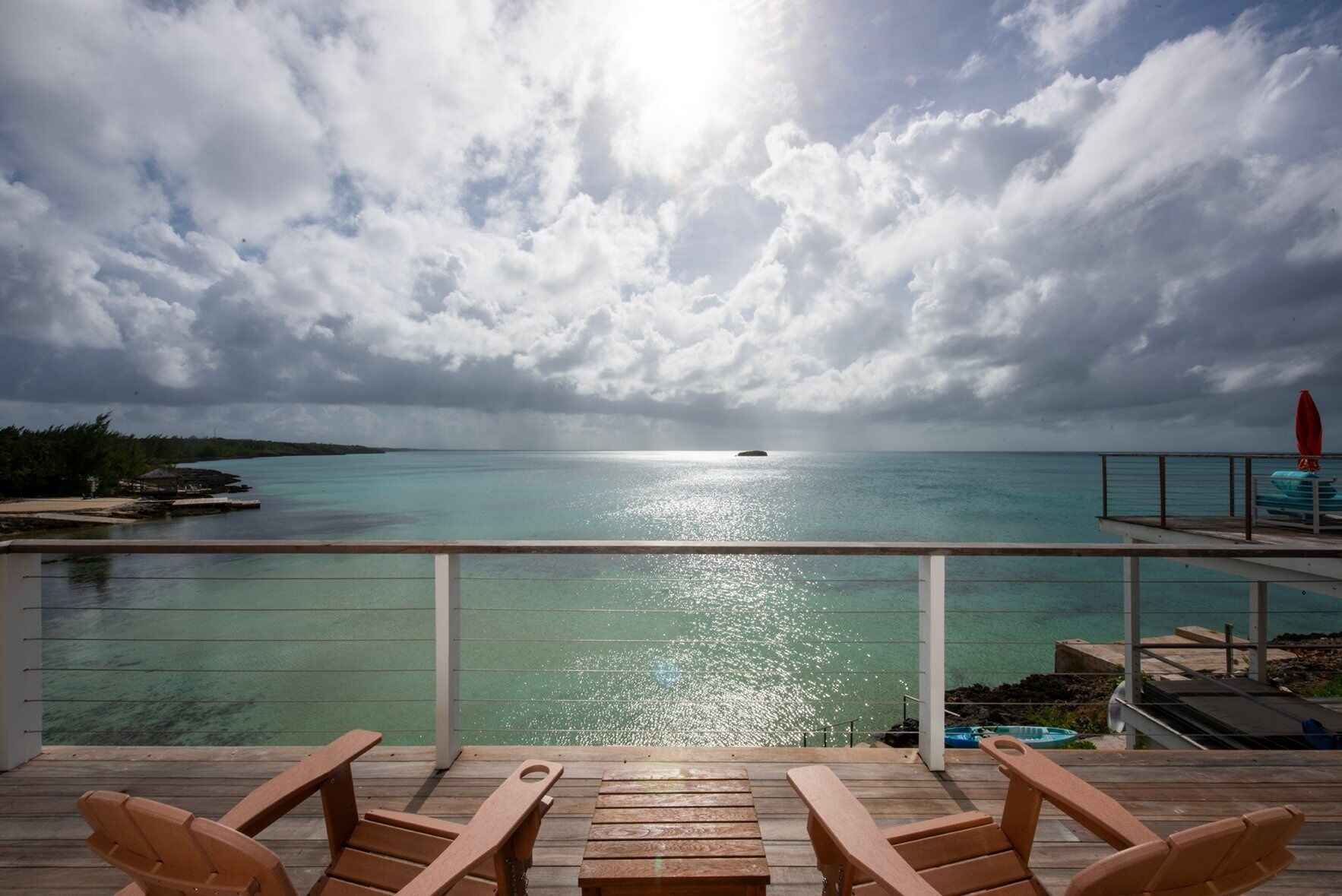 A deck with wooden chairs overlooking turquoise waters, a distant island, and a cloudy sky.