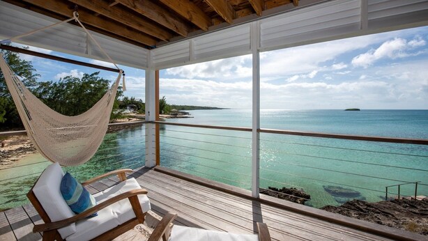 A wooden deck with a hammock chair, clear turquoise waters, and a view of a distant island. - Touch Of Class Cottage - A Caribbean Beachfront Cottage 2 Bedroom Cottage (Savannah Sound)