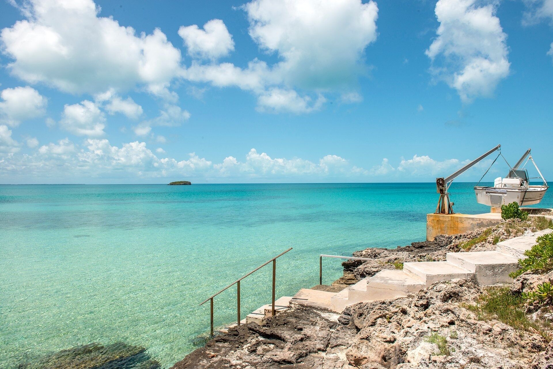 A coastal scene with clear turquoise waters, a small island in the distance, and a boat docked on a rocky shore with steps leading down to the water.