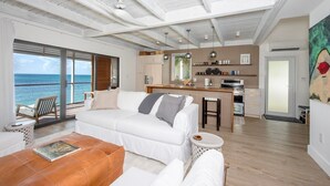 A spacious living area with a white sofa, a coffee table, and a view of the ocean. - Touch Of Class Cottage - A Caribbean Beachfront Cottage 2 Bedroom Cottage (Savannah Sound)