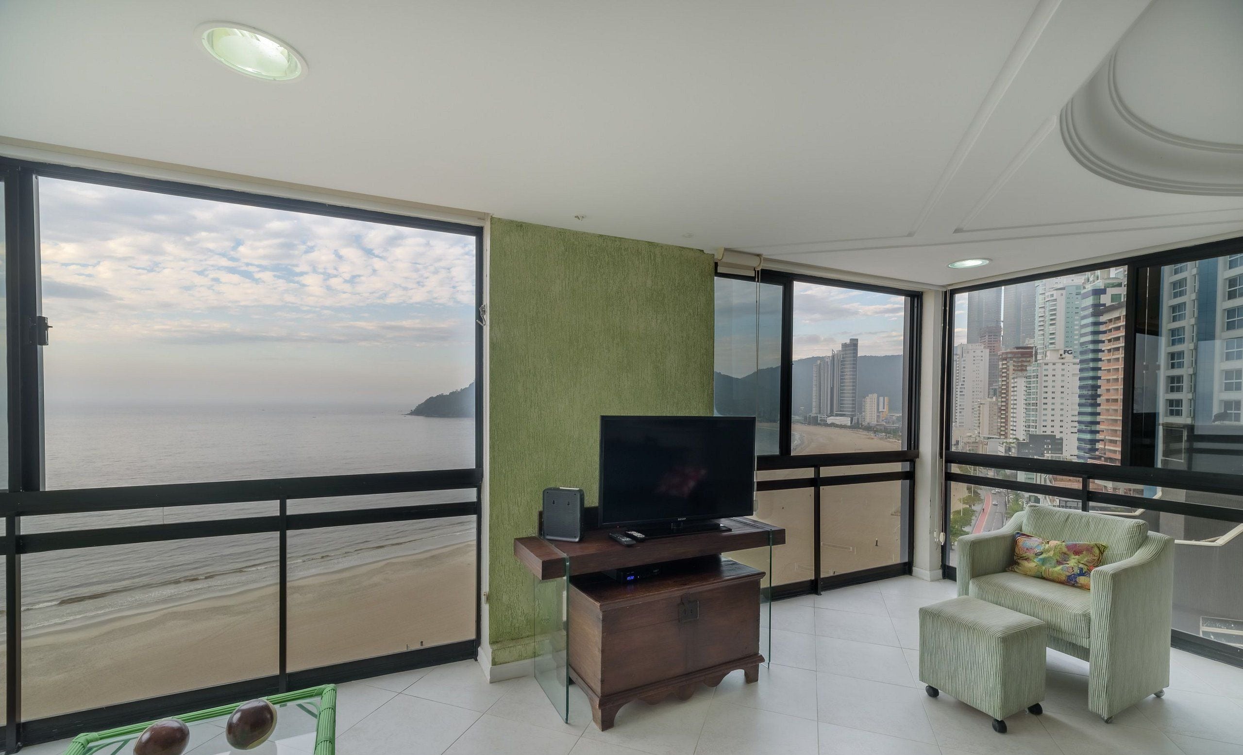 Un salón amplio con televisor grande, sofá y vistas al océano y al paisaje urbano a través de grandes ventanales.