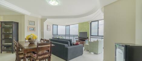 A spacious living room with a dining area, a sofa set, a television, and a large window offering a view.