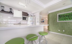 A modern kitchen with white cabinets, a microwave, a refrigerator with magnets, and a green wall with a framed picture. There are three green stools at the counter. - 2BR Seafront apt W/garage in BC Nvi501 (Balneário Camboriú)