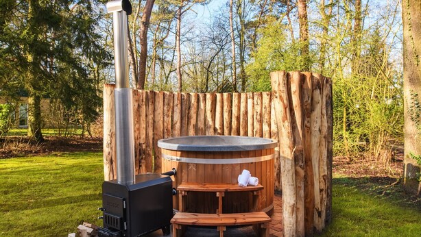 Een houtgestookte sauna met een bank en handdoek, omringd door een houten omheining in een bebost gebied.