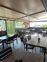 Área de terraço ao ar livre com mesas, cadeiras, plantas em vasos e vista para um edifício de madeira ao fundo.