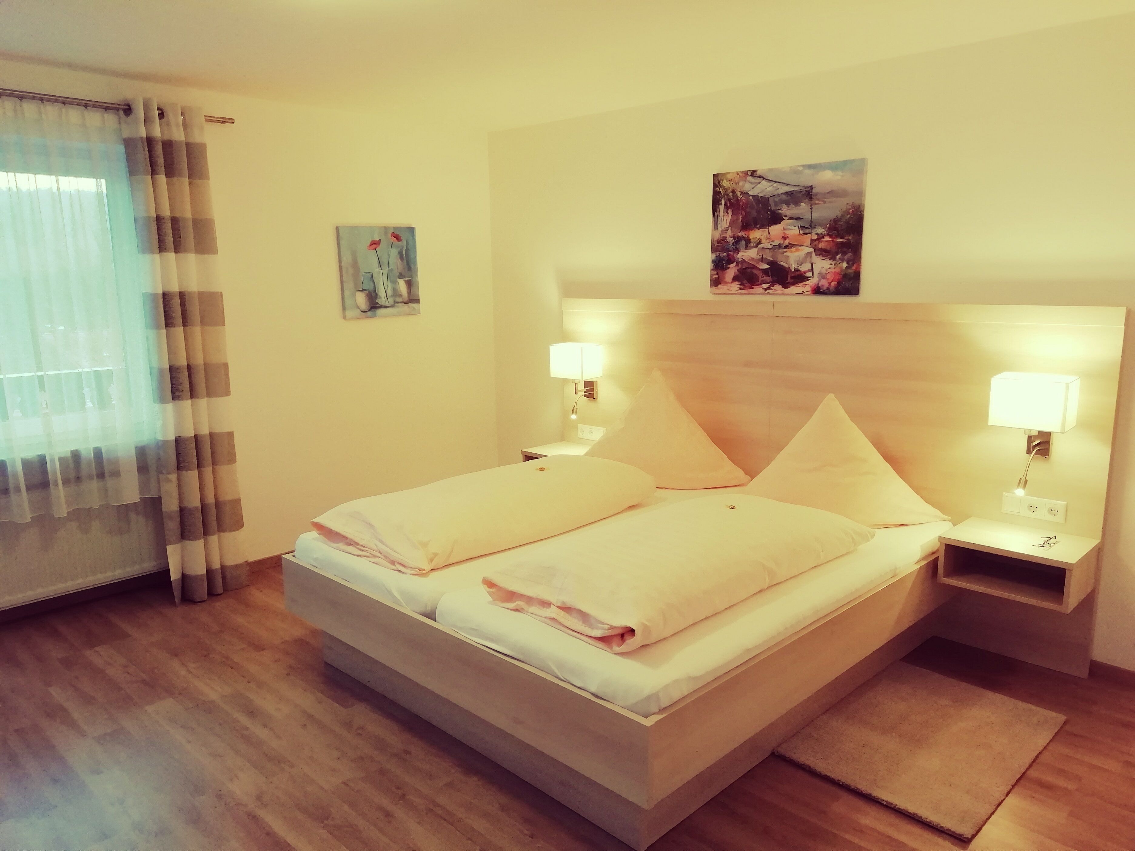 A hotel room with a bed, two bedside lamps, a painting above the bed, and a window with curtains.