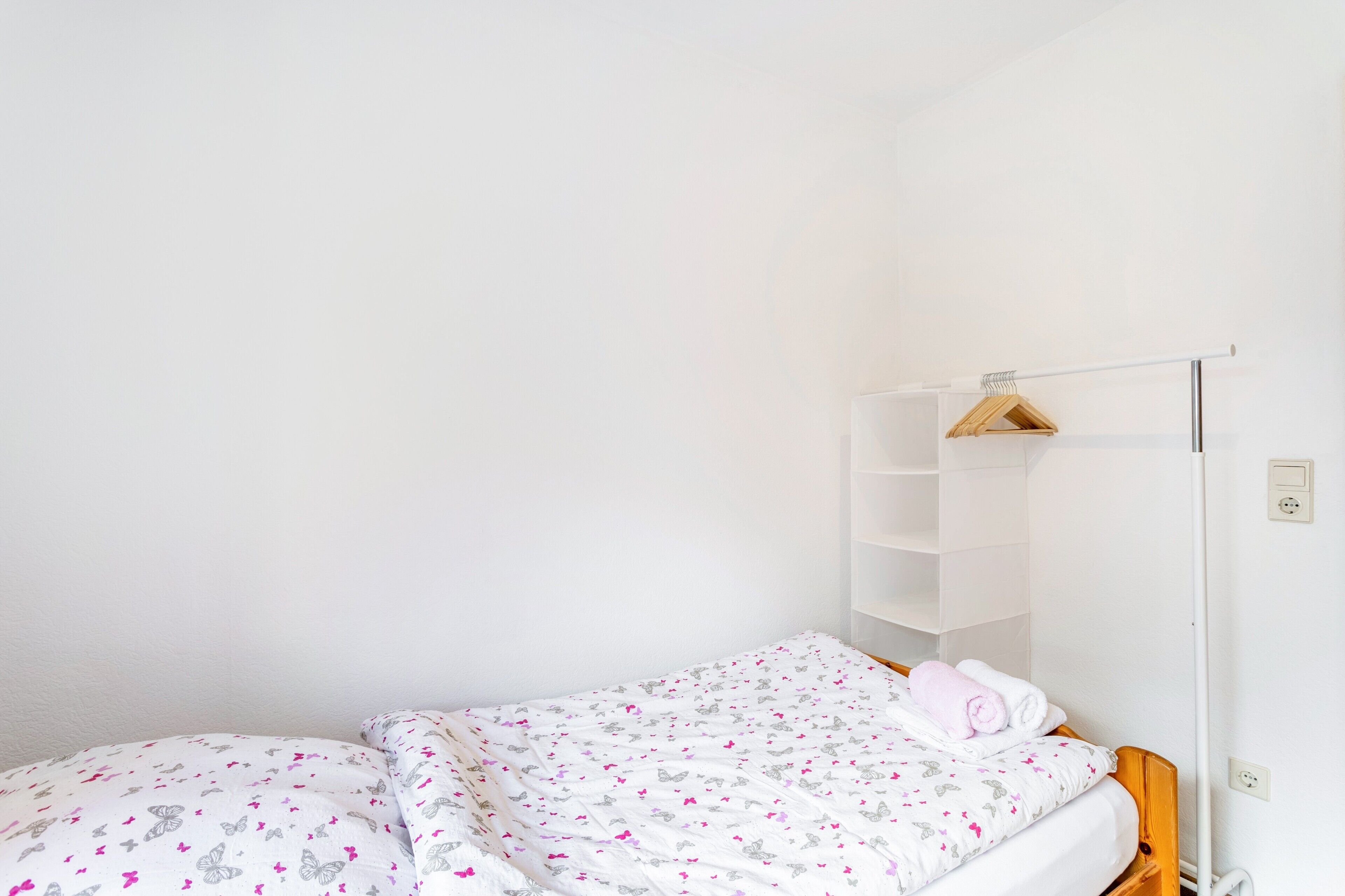 A single bed with a patterned bedspread, a wooden headboard, and a white wall with a shelf unit.