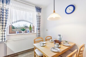 Un comedor con una mesa dispuesta para dos personas, sillas, una lámpara colgante, un reloj en la pared y una ventana con cortinas.