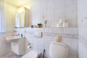 Baño con lavamanos, espejo, inodoro y repisas con artículos de aseo.