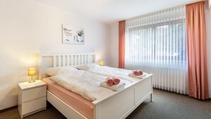 Uma cama de casal com roupa de cama branca, uma mesa de cabeceira com abajur, um quadro na parede e cortinas leves com uma faixa peachy.