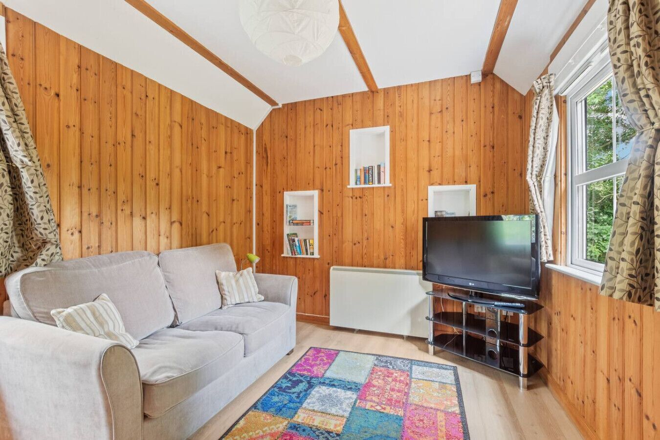 A cozy living room with a sofa, a flat-screen TV, a bookshelf, and a window with curtains.