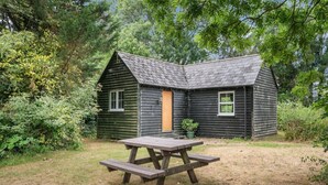 Une petite cabane en bois sombre, avec un toit en pente, une seule porte et deux fenêtres. Une table de pique-nique se trouve devant la cabane.
