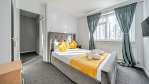Ein modernes Hotelzimmer mit grau gestepptem Kopfteil, einem Bett mit weißer und gelber Bettwäsche, einem Nachttisch aus Holz und einem Fenster mit grünen Vorhängen.