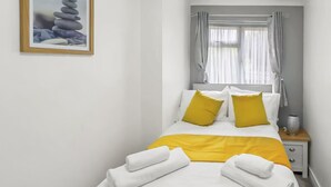 Un lit bien fait, avec une literie blanche et jaune, une table de chevet avec une lampe et un tableau encadré au mur.