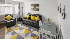 Un salon moderne avec un canapé en cuir noir, des coussins jaunes, un tapis à motifs géométriques, une table à manger avec des chaises et un tableau accroché au mur.