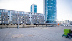 A waterfront promenade with modern buildings, a clear sky, and a green cart with crates.