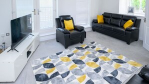 A living room with a flat-screen TV on a white stand, two black leather sofas with yellow cushions, a patterned rug, and a window with blinds.